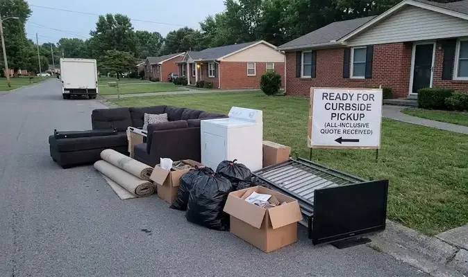 A pile of household items, including a sectional couch, a dryer, carpets, boxes, trash bags, a TV, and a mattress frame, sits on the street curb in front of a brick house. A sign reads, 'READY FOR CURBSIDE PICKUP (ALL-INCLUSIVE QUOTE RECEIVED).' A white truck is visible in the background on the left.