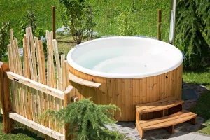 Wooden hot tub in a lush garden, surrounded by tall grasses and a rustic wooden fence. Wooden steps lead to the tub, creating a serene, natural vibe.