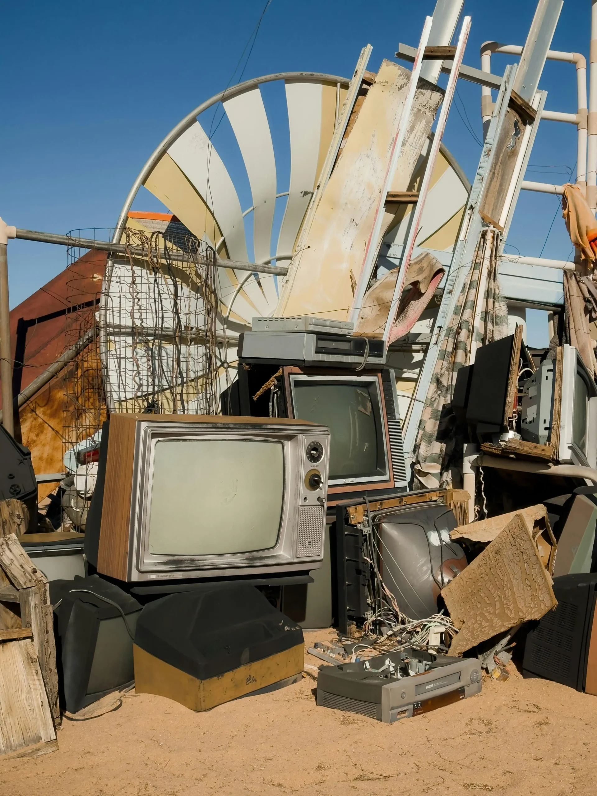 A clutter of vintage electronics and debris against a clear blue sky. Old TVs and various scrap items are haphazardly stacked, evoking nostalgia and decay.
