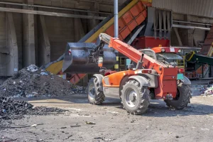 An orange loader vehicle in an industrial area, surrounded by piles of debris and machinery, conveying a busy and gritty work environment.