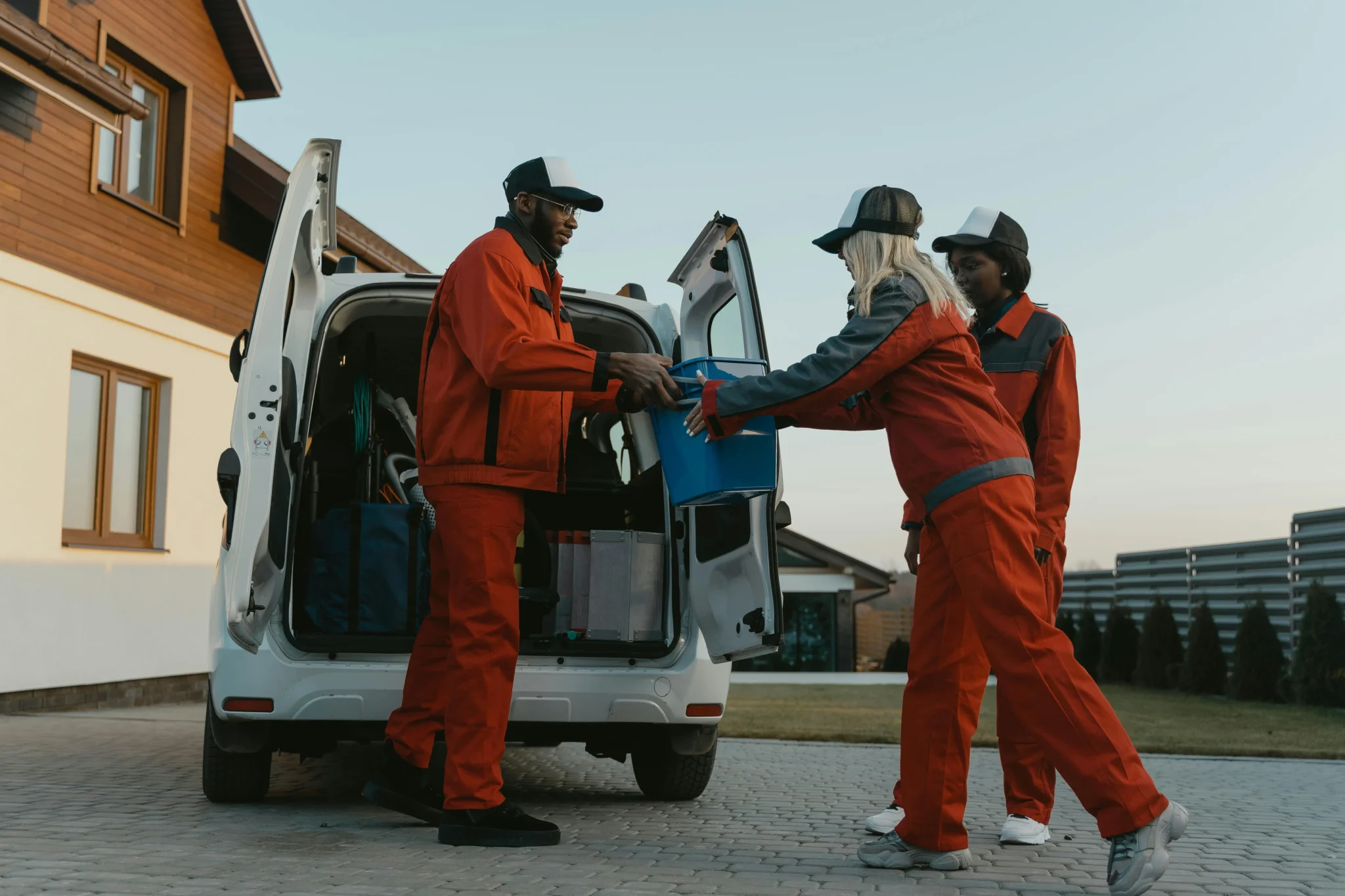 Three people in red uniforms load a blue bin from a white van parked beside a modern house. The scene conveys teamwork and efficiency.